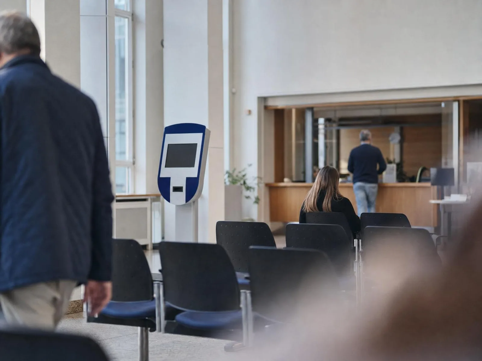 Salle d'attente d'un service municipal allemand au petit matin : rangées de chaises vides, distributeur de tickets numérotés et comptoir en bois flou à l'arrière-plan, lumière douce venant de hautes fenêtres.