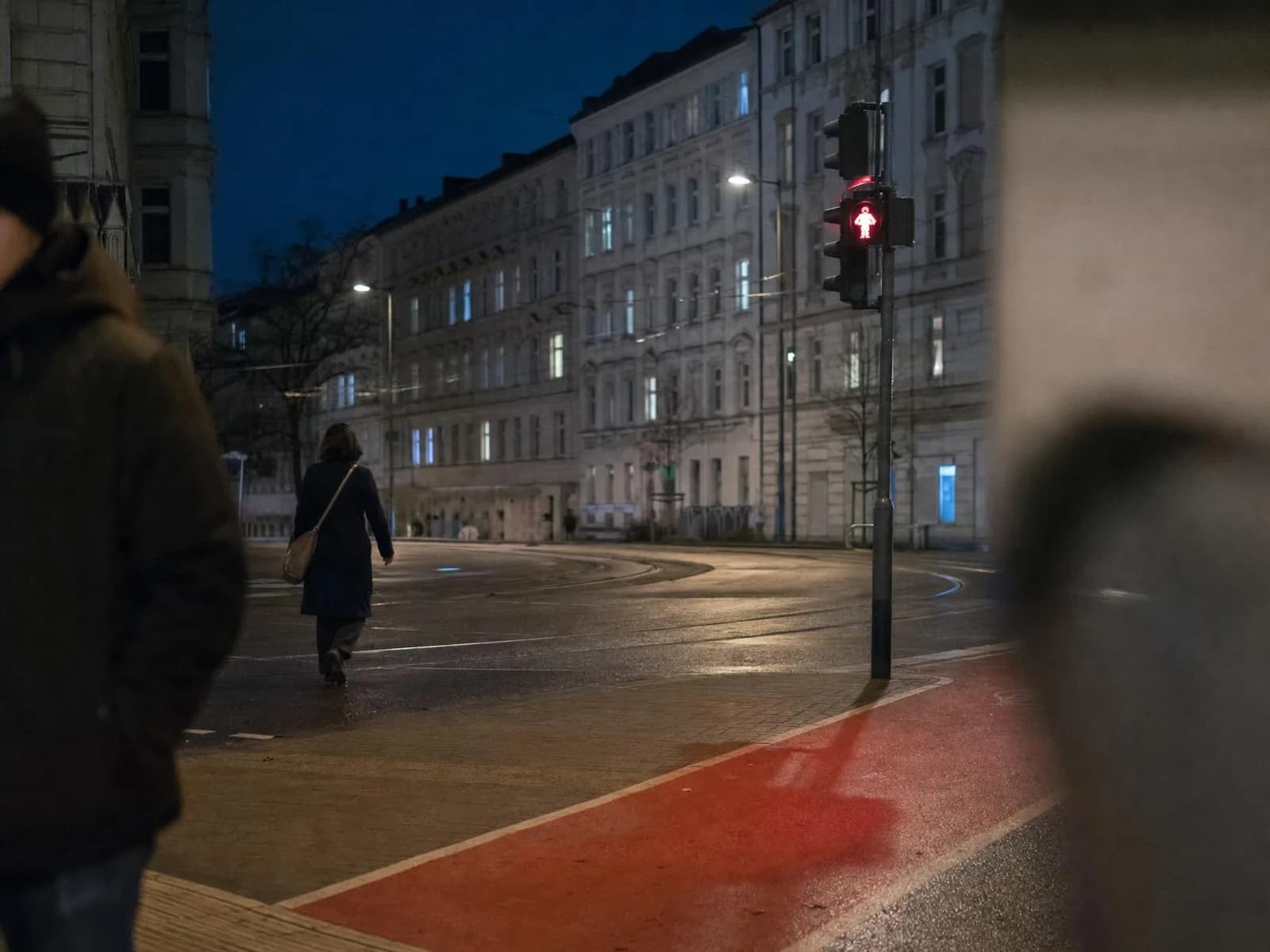Un large carrefour urbain désert la nuit, asphalte mouillé reflétant la lueur rouge d'un signal piéton, rails de tramway et façades d'immeubles anciens aux fenêtres éclairées à l'arrière-plan.