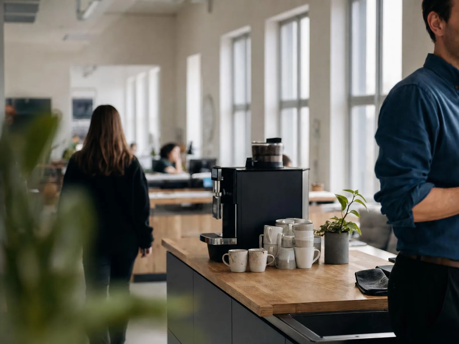 Un coin cuisine d'entreprise baigné de lumière : machine à café sur un plan de travail en bois, deux tasses posées, quelques mugs empilés, des silhouettes floues au fond de la pièce.