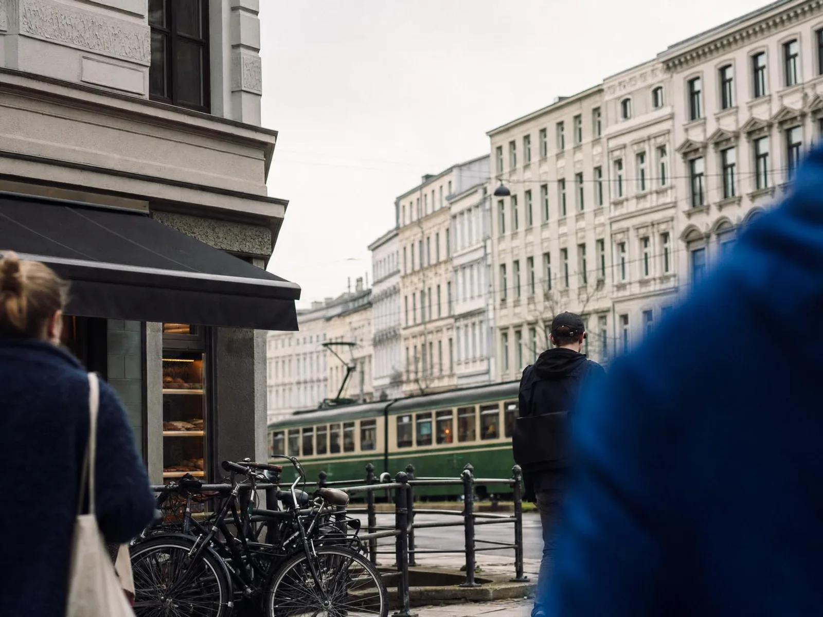 Rue résidentielle allemande en fin de matinée : façades anciennes à hautes fenêtres, vélos alignés le long d'une grille, un tram vert qui passe au fond sous une lumière grise et douce.