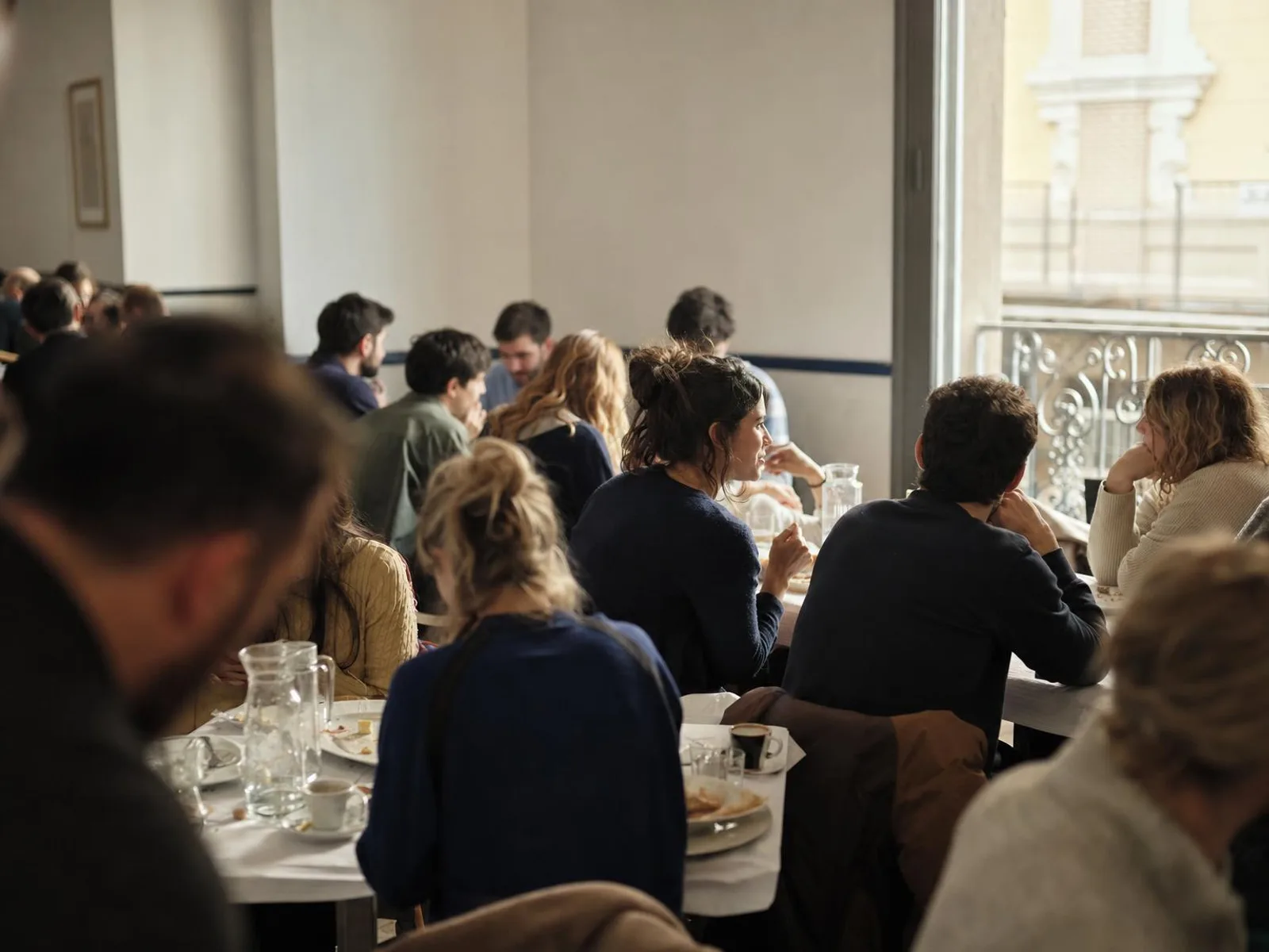 Salle d'un restaurant de quartier madrilène en milieu d'après-midi : tables encore garnies, tasses de café, lumière chaude venant d'une fenêtre ouverte sur une façade de pierre claire.