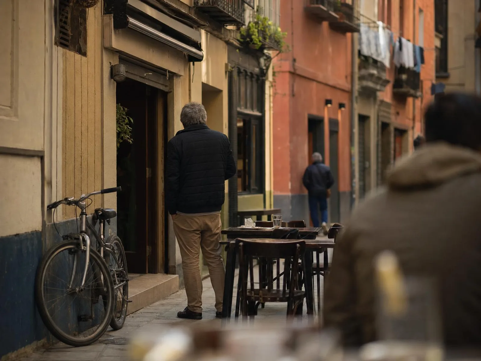 Rue étroite d'un centre-ville espagnol en fin d'après-midi, façades ocre, balcons en fer forgé et petites tables de bar installées sur le trottoir devant une porte ouverte.