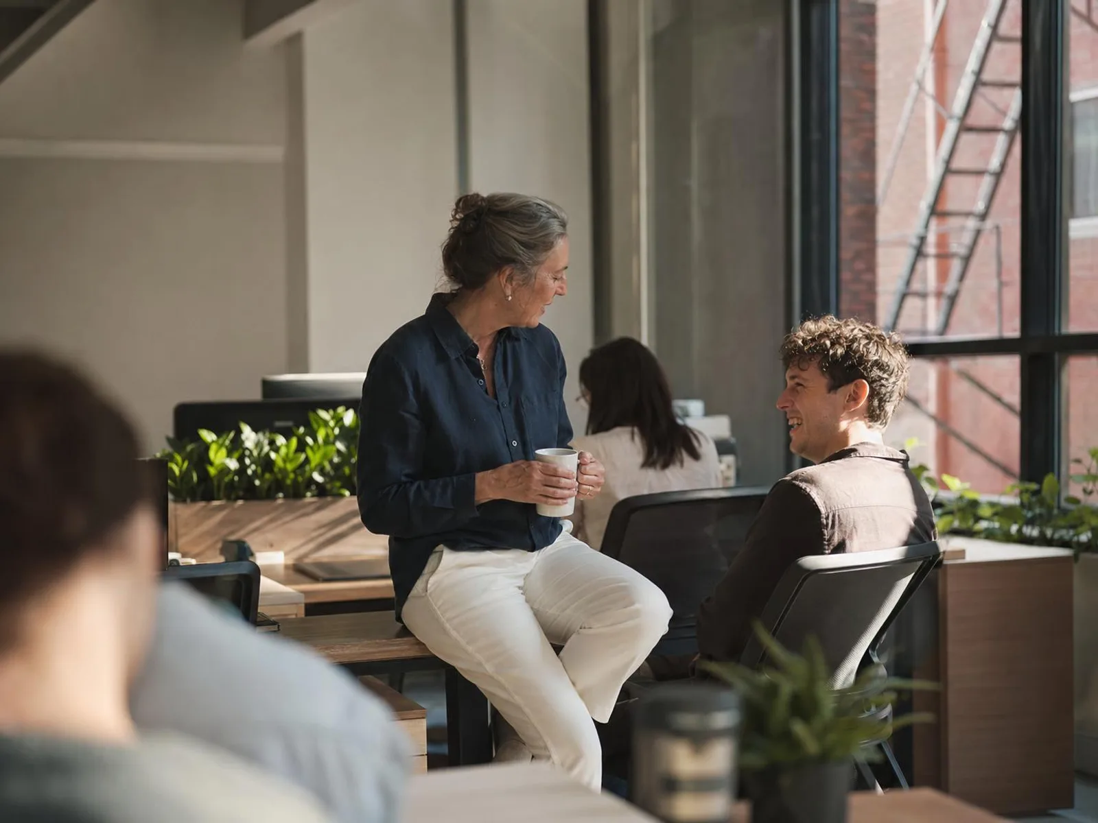 Dans un bureau ouvert, une femme d'une cinquantaine d'années s'est assise au bord du bureau d'un collègue plus jeune, un café à la main ; il a fait pivoter sa chaise vers elle et rit.
