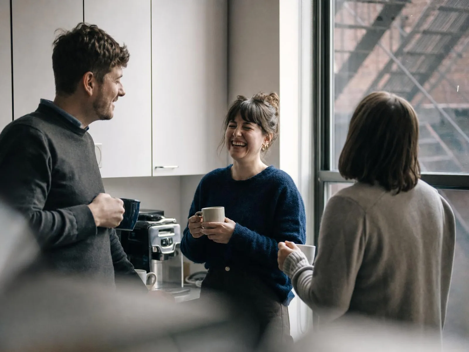 Trois collègues debout dans un coin cuisine de bureau, tasses à la main, en pleine conversation ; l'une d'elles rit, une autre est de dos.