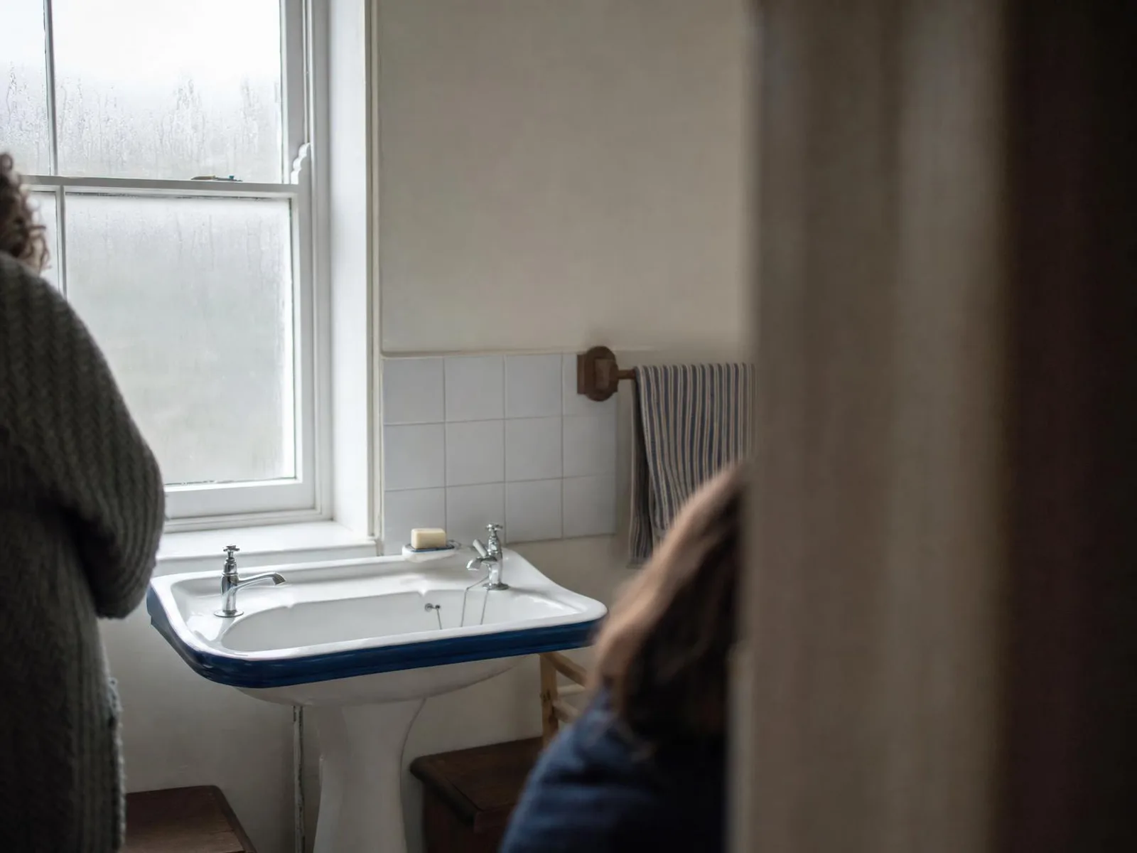 Un lavabo en porcelaine blanche à liseré bleu, avec deux robinets chromés placés à chaque extrémité, dans une salle de bains ancienne éclairée par une petite fenêtre givrée.
