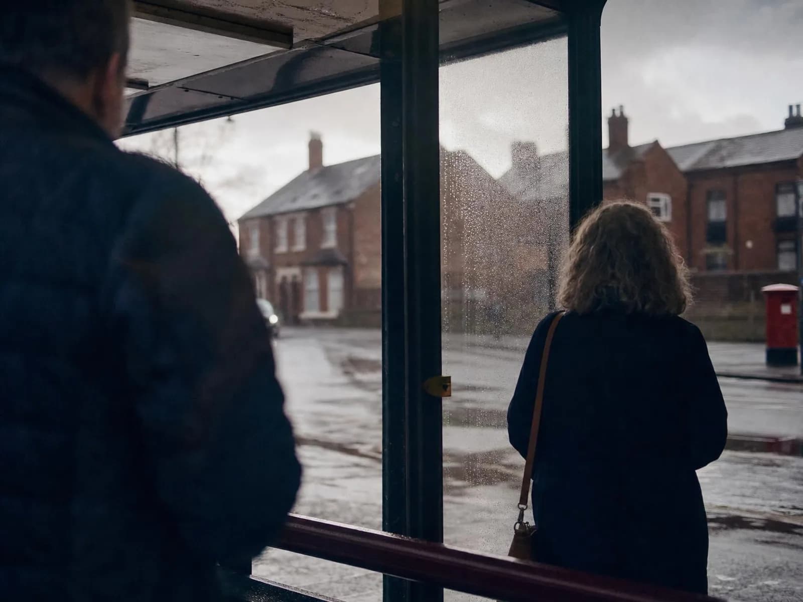 Un abribus de rue britannique après l'averse, deux silhouettes floues sous le auvent, des maisons de brique en face et le ciel gris qui s'éclaircit dans les flaques.