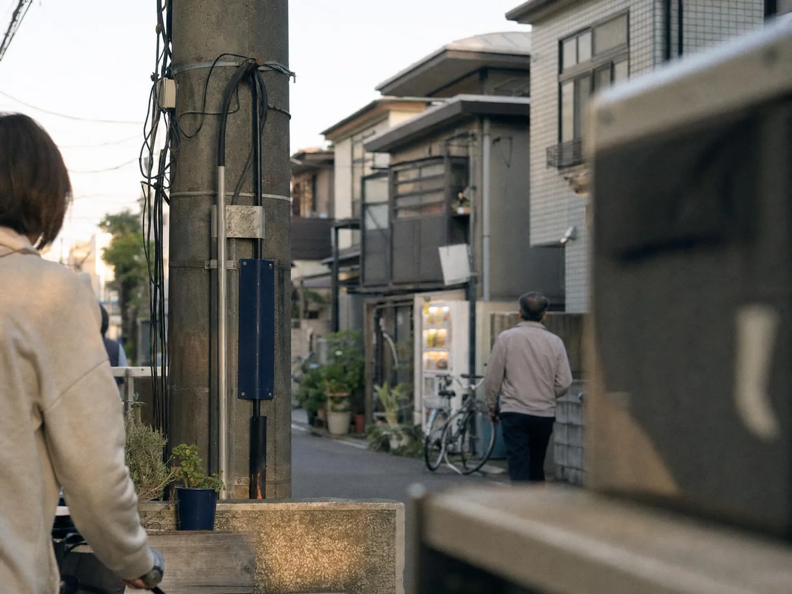 Ruelle résidentielle japonaise en fin de journée : poteau électrique et câbles au premier plan, maisons basses avec plantes en pots, vélos appuyés contre un mur et distributeur de boissons éclairé au fond.