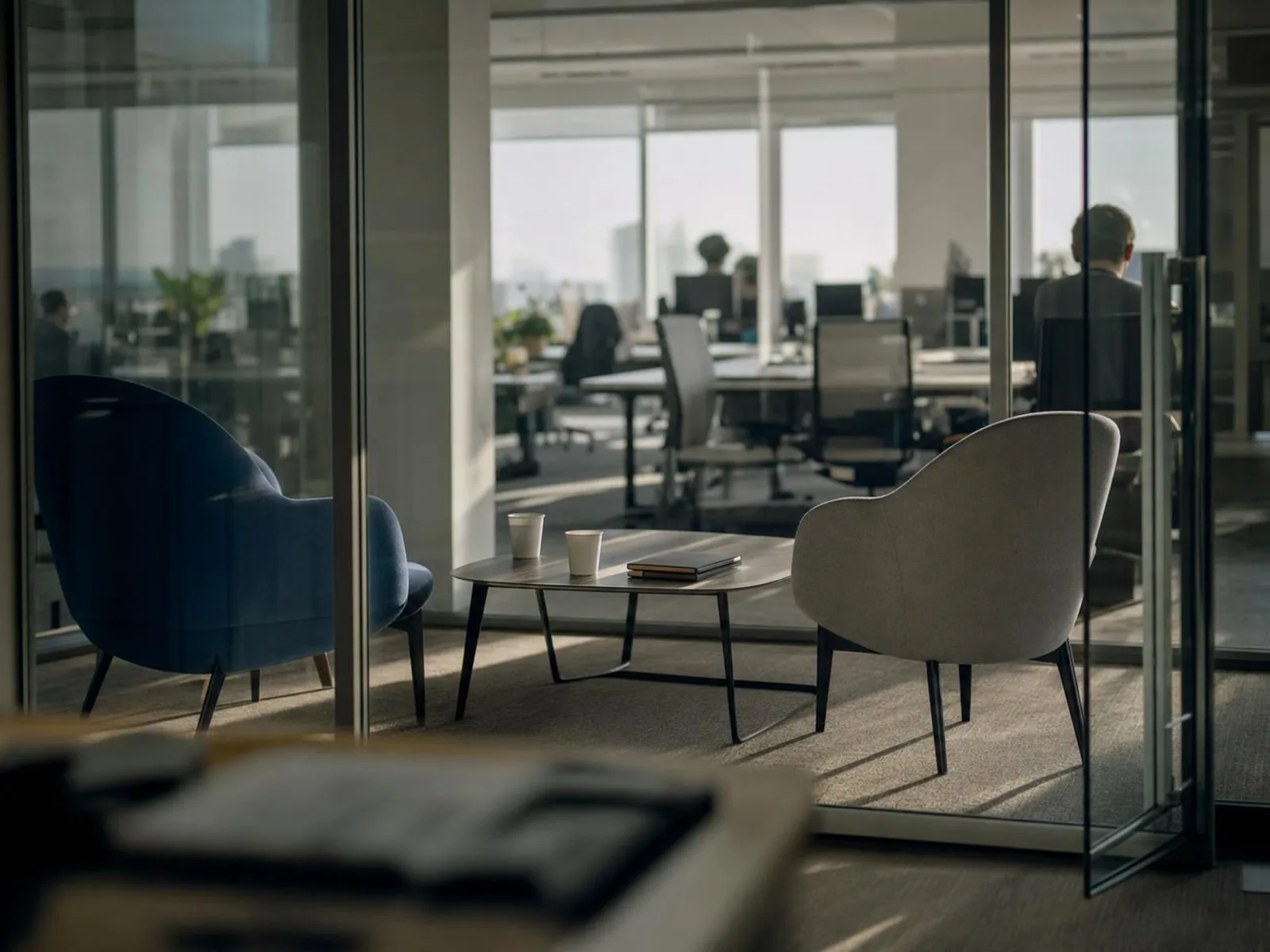 Une petite salle de réunion vitrée dans un bureau en open space, deux chaises face à face autour d'une table basse avec deux gobelets et un carnet fermé, dans la lumière de fin d'après-midi.