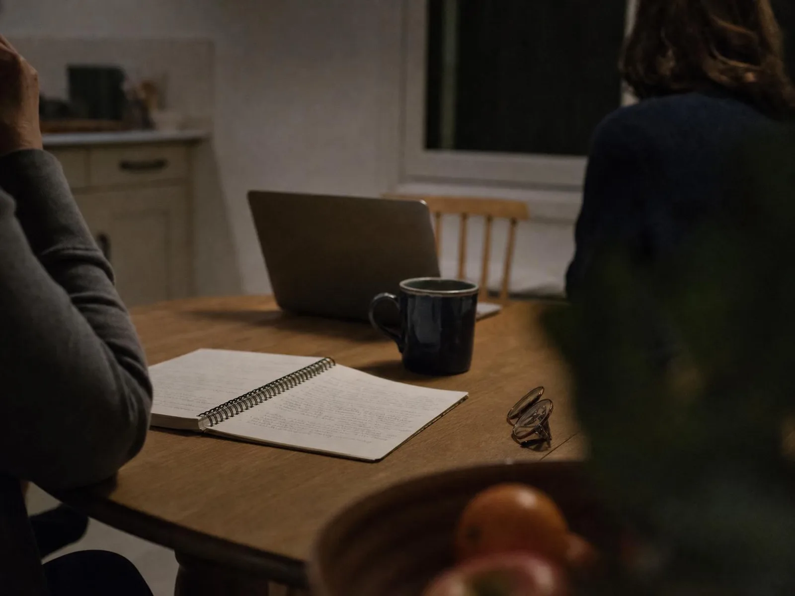 Table de cuisine le soir, sous une lampe chaude : un carnet ouvert couvert de notes manuscrites, une tasse de thé, une paire de lunettes posée à côté, un ordinateur portable entrouvert au fond.