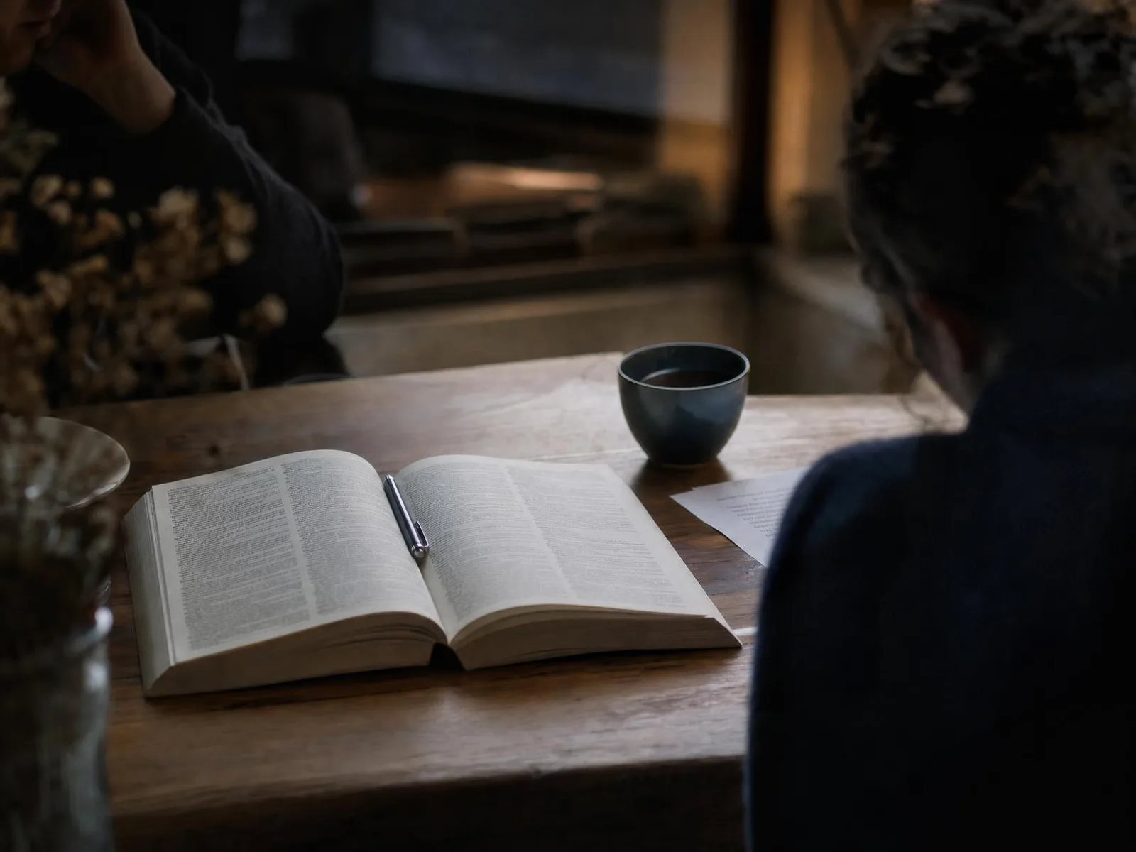 Sur un bureau en bois éclairé par une lampe, un dictionnaire ouvert et des pages imprimées annotées, un stylo posé en travers et une tasse de thé un peu à l'écart.