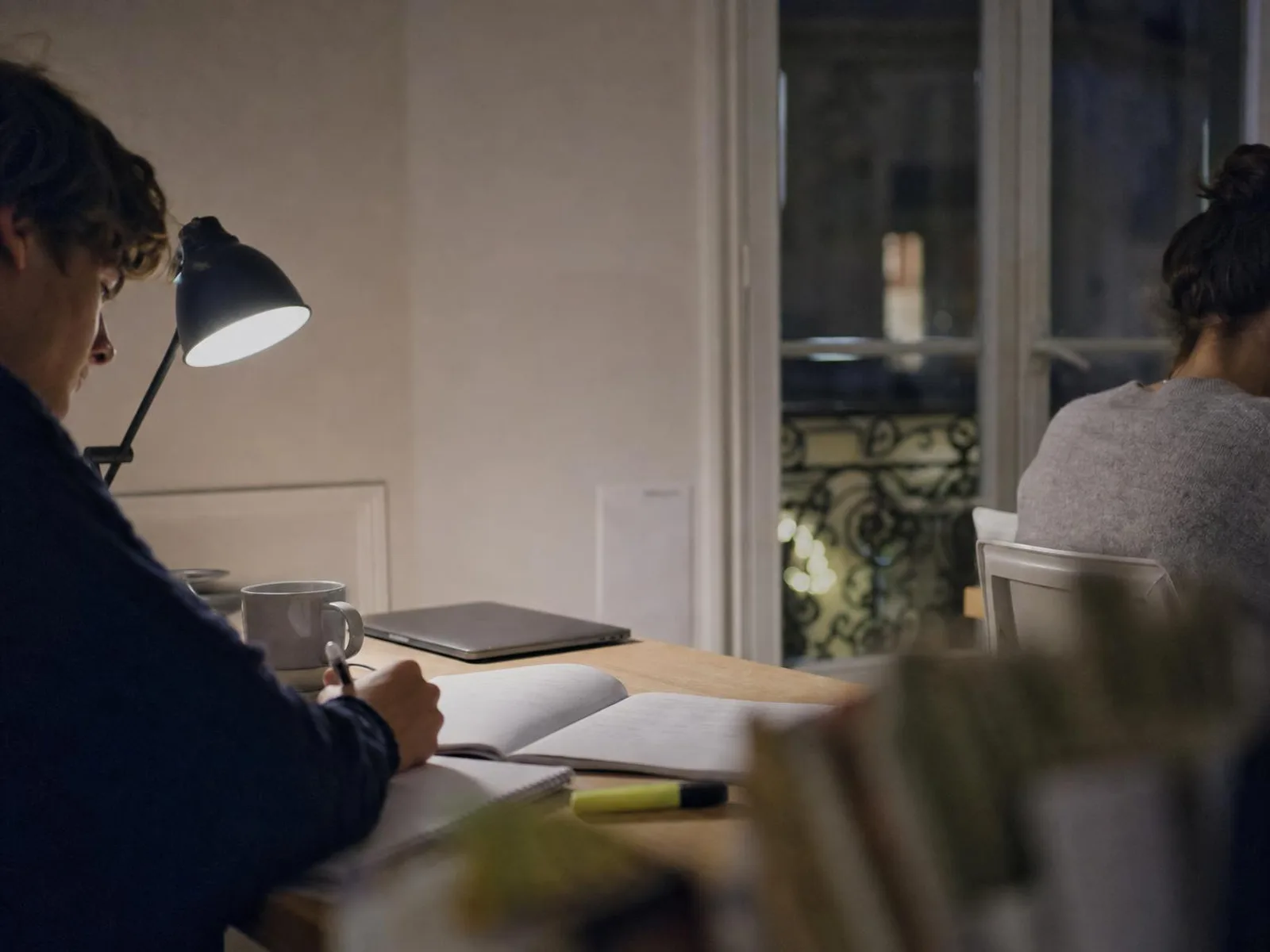 Un coin de bureau éclairé par une lampe, un cahier ouvert et un surligneur posés sur le bois, une tasse de thé, et derrière la fenêtre la nuit sur une façade parisienne.