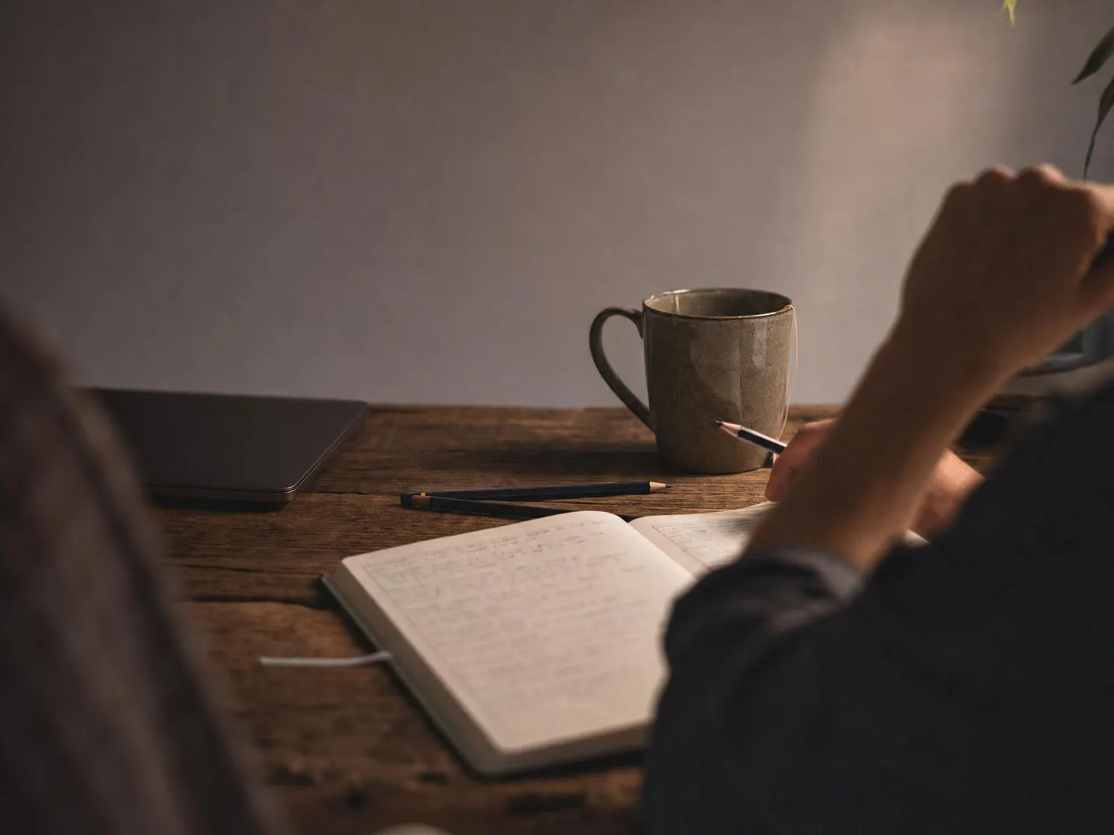 Un bureau en bois éclairé par une lampe chaude, un cahier ouvert couvert de notes manuscrites, deux crayons et une tasse de thé, un ordinateur portable fermé sur le côté.
