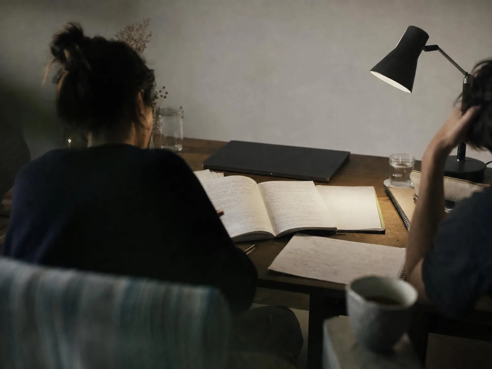 Bureau éclairé par une lampe la nuit, un carnet ouvert couvert de notes manuscrites avec des flèches tracées entre les paragraphes, un ordinateur portable de dos et une tasse de thé au bord du cadre.
