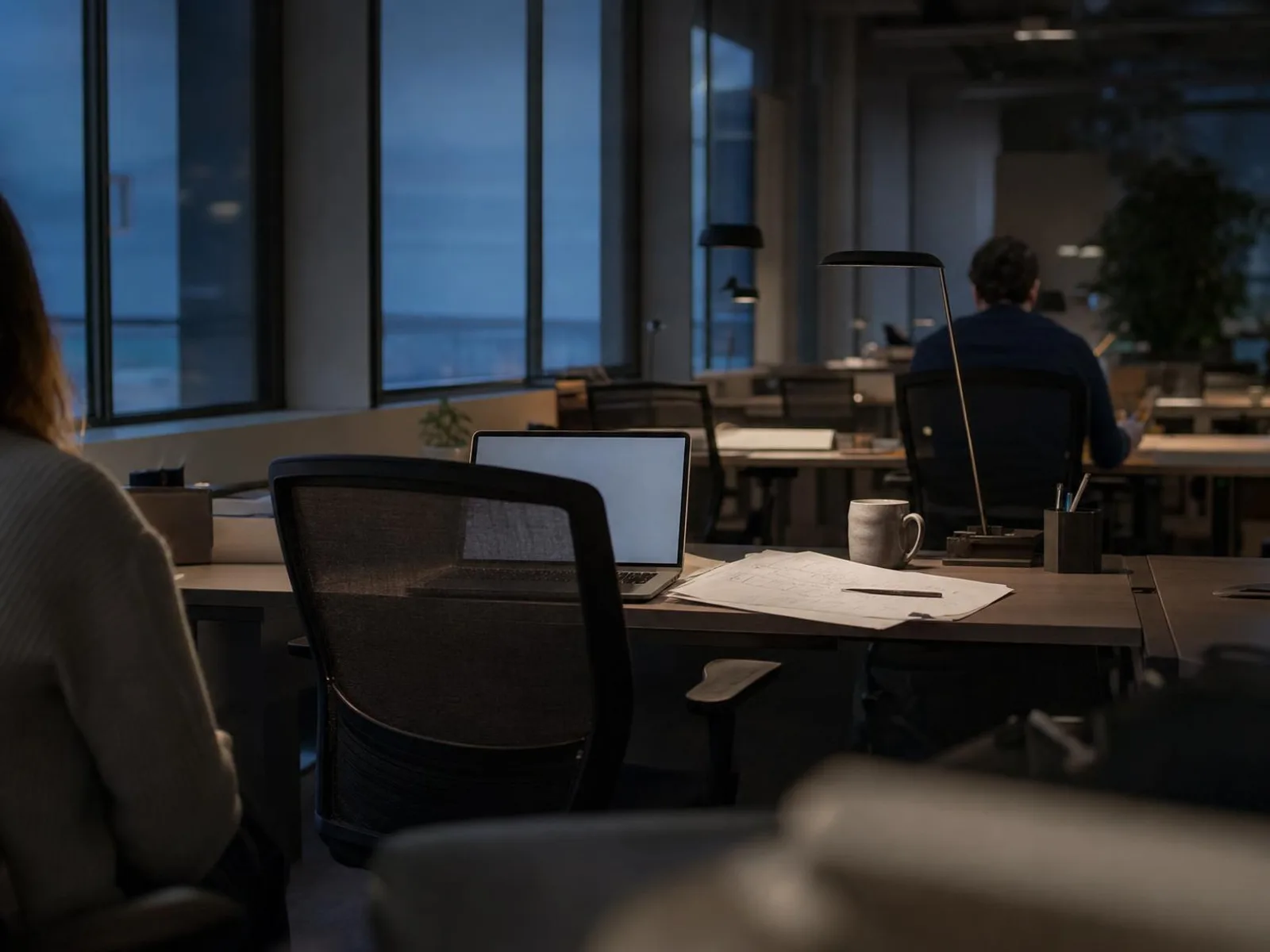 Un bureau en fin de journée, éclairé par une lampe d'appoint : un ordinateur portable ouvert, un document imprimé annoté au crayon et une tasse posée à côté, devant une baie vitrée où tombe le soir.