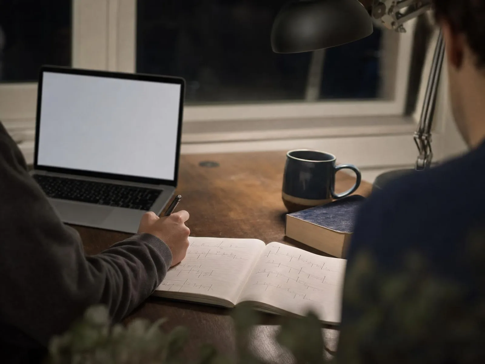 Un coin de bureau éclairé par une lampe, un cahier ouvert couvert de notes manuscrites, un ordinateur portable, une tasse et un dictionnaire posé à plat.
