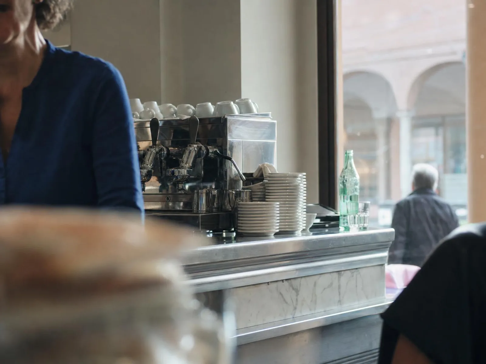 Comptoir en marbre d'un bar italien au petit matin : machine à expresso chromée, tasses retournées, une bouteille d'eau pétillante et un verre, lumière rasante venue de la rue à arcades.