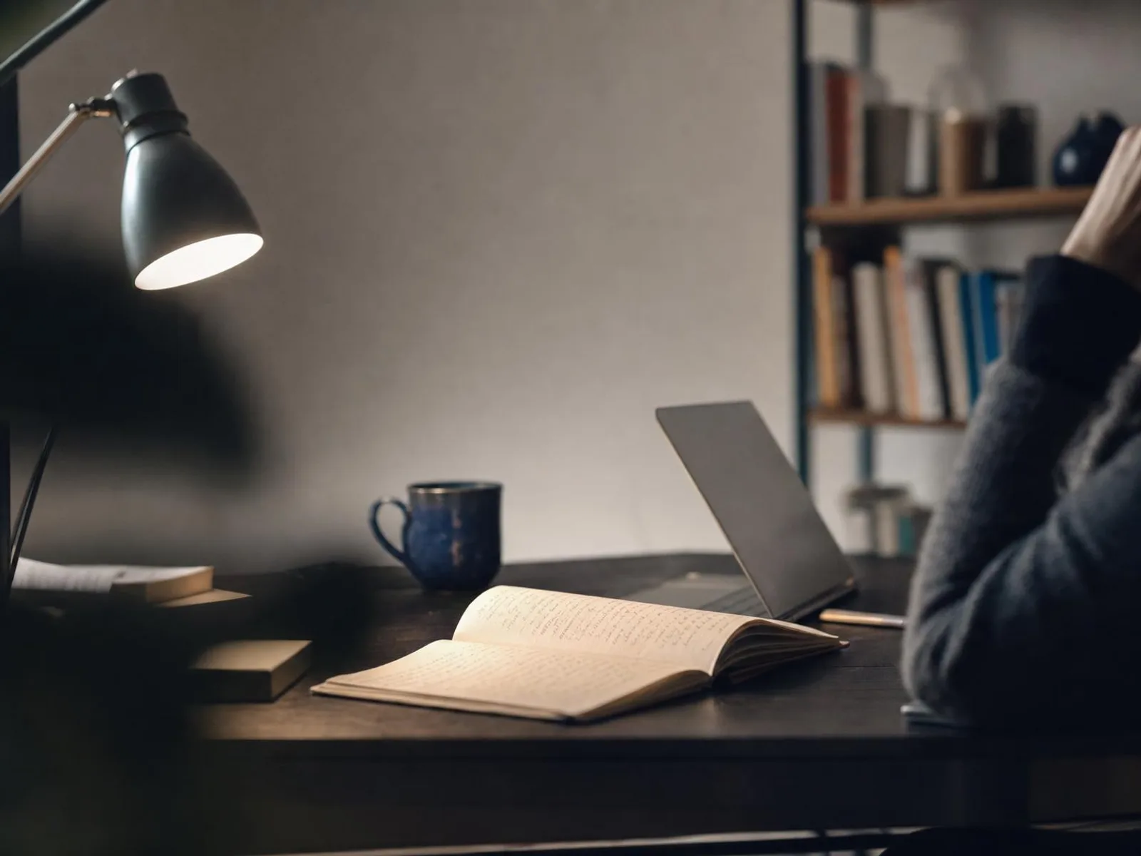 Un bureau éclairé par une lampe le soir, un cahier ouvert couvert de notes manuscrites, une tasse de thé et un ordinateur portable entrouvert, des étagères de livres floues à l'arrière-plan.