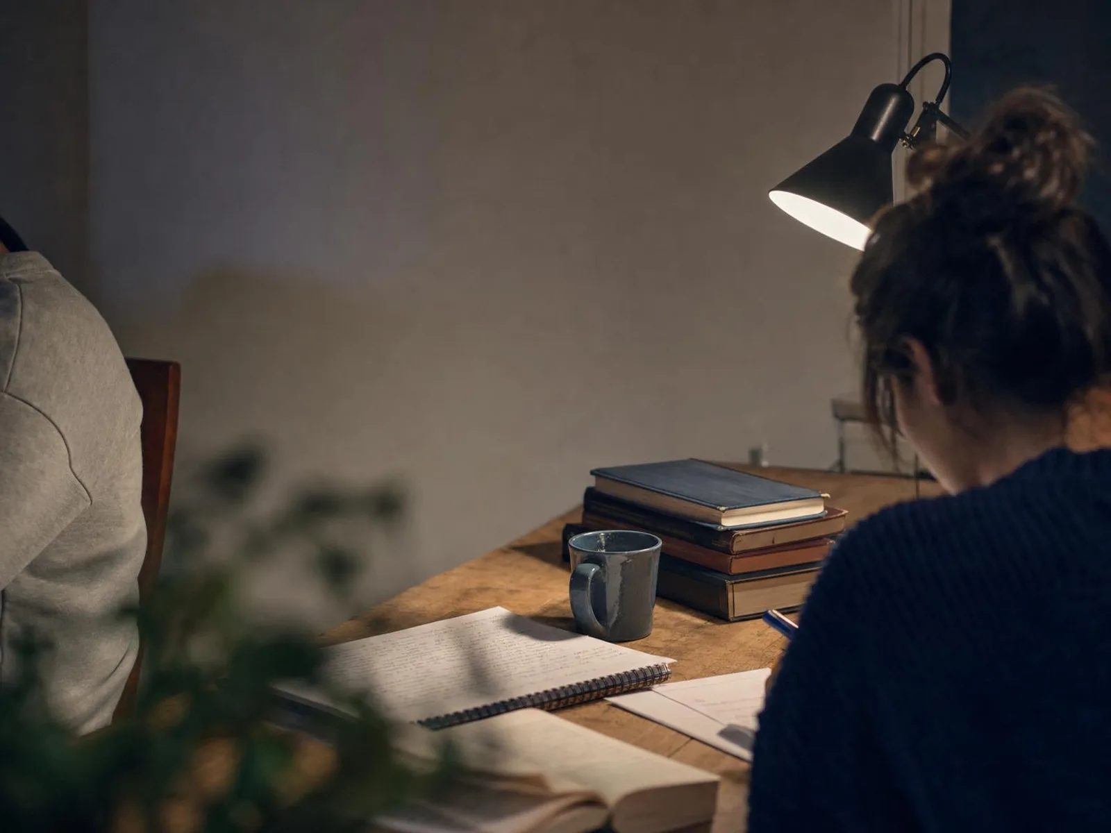 Bureau éclairé par une lampe la nuit, cahier ouvert couvert de notes manuscrites, quelques ouvrages de référence empilés et une tasse posée à côté.