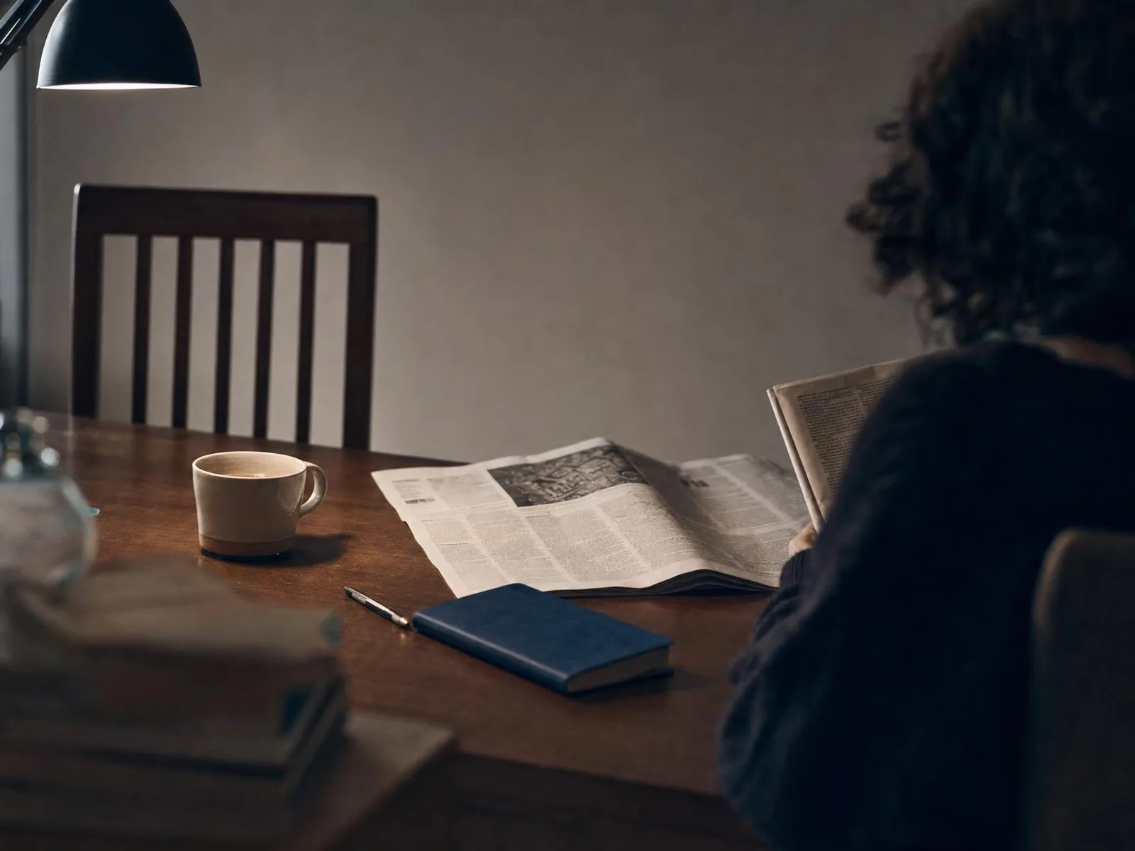 Un bureau éclairé par une lampe, un journal ouvert à plat, un carnet fermé, un crayon et une tasse de thé à moitié bue.