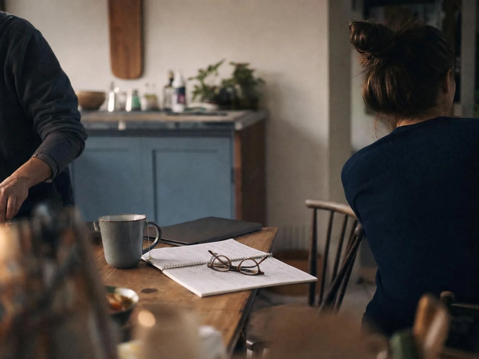 Une table de cuisine le soir, éclairée par une lampe de côté : un cahier à spirale ouvert, une tasse de thé, des lunettes posées sur la page et un ordinateur portable entrouvert derrière.