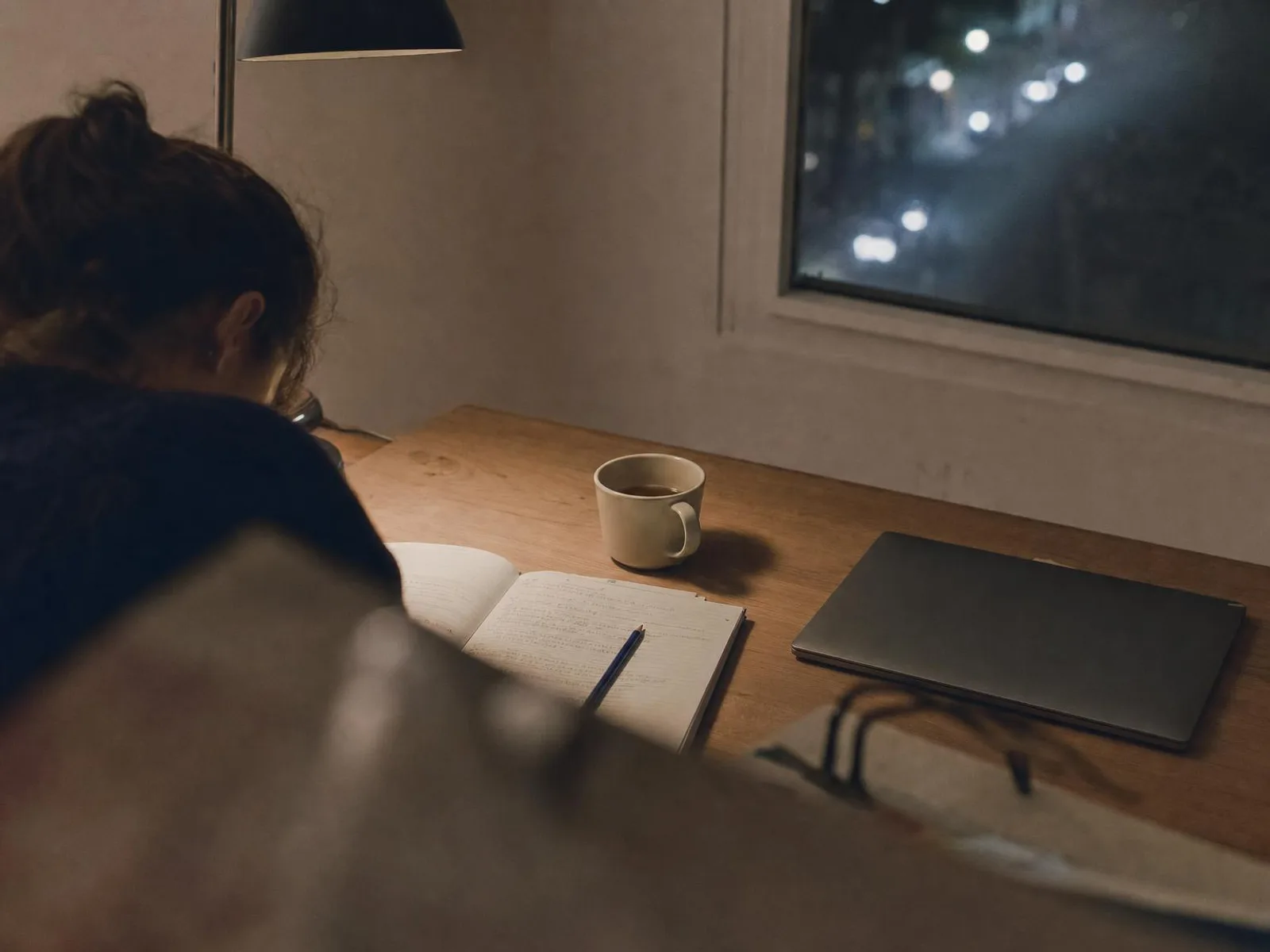 Un bureau en bois éclairé par une lampe, la nuit : un cahier ouvert couvert d'écriture manuscrite, un crayon, une tasse de thé à moitié vide, et une fenêtre sombre au fond.