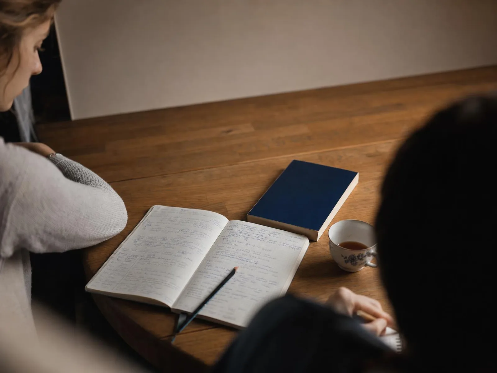 Bureau en bois éclairé par une lampe, la nuit : un cahier ouvert couvert de notes manuscrites, un dictionnaire de poche posé à plat, une tasse de thé et un crayon en travers de la page.