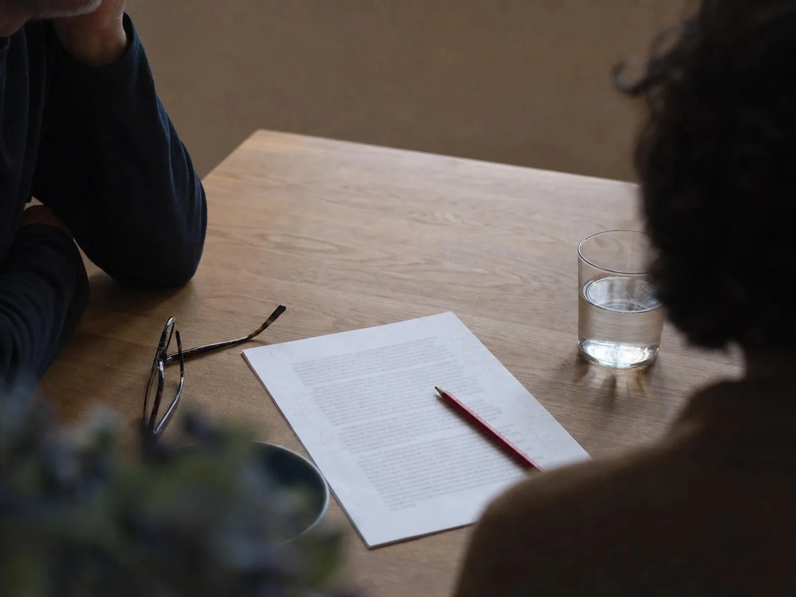Bureau en bois éclairé par une lampe, une page imprimée couverte d'annotations au crayon, un crayon rouge posé en travers, des lunettes pliées sur le bord de la feuille.
