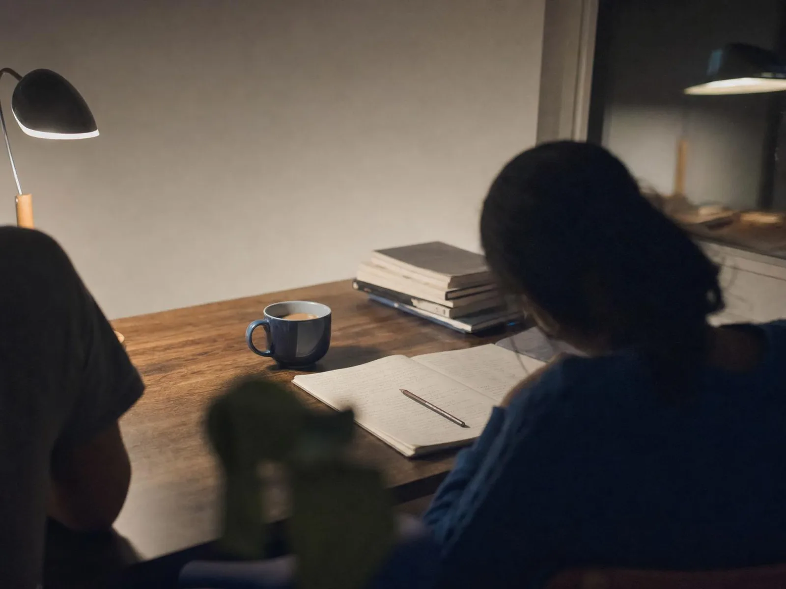 Une table de travail éclairée par une lampe le soir, un cahier ouvert couvert d'écriture, un crayon posé en travers, une tasse et quelques livres empilés.