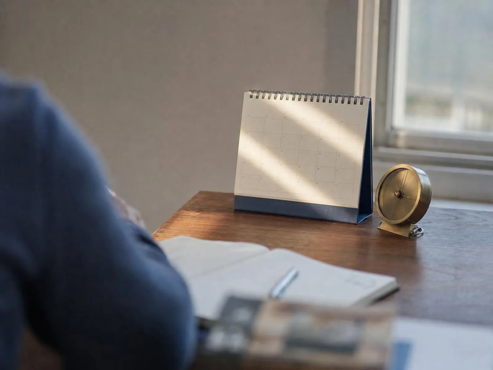 Sur un bureau en bois près d'une fenêtre, un calendrier mural et une petite pendule de cuivre côte à côte, baignés d'une lumière rasante de fin d'après-midi, avec un carnet ouvert au premier plan.