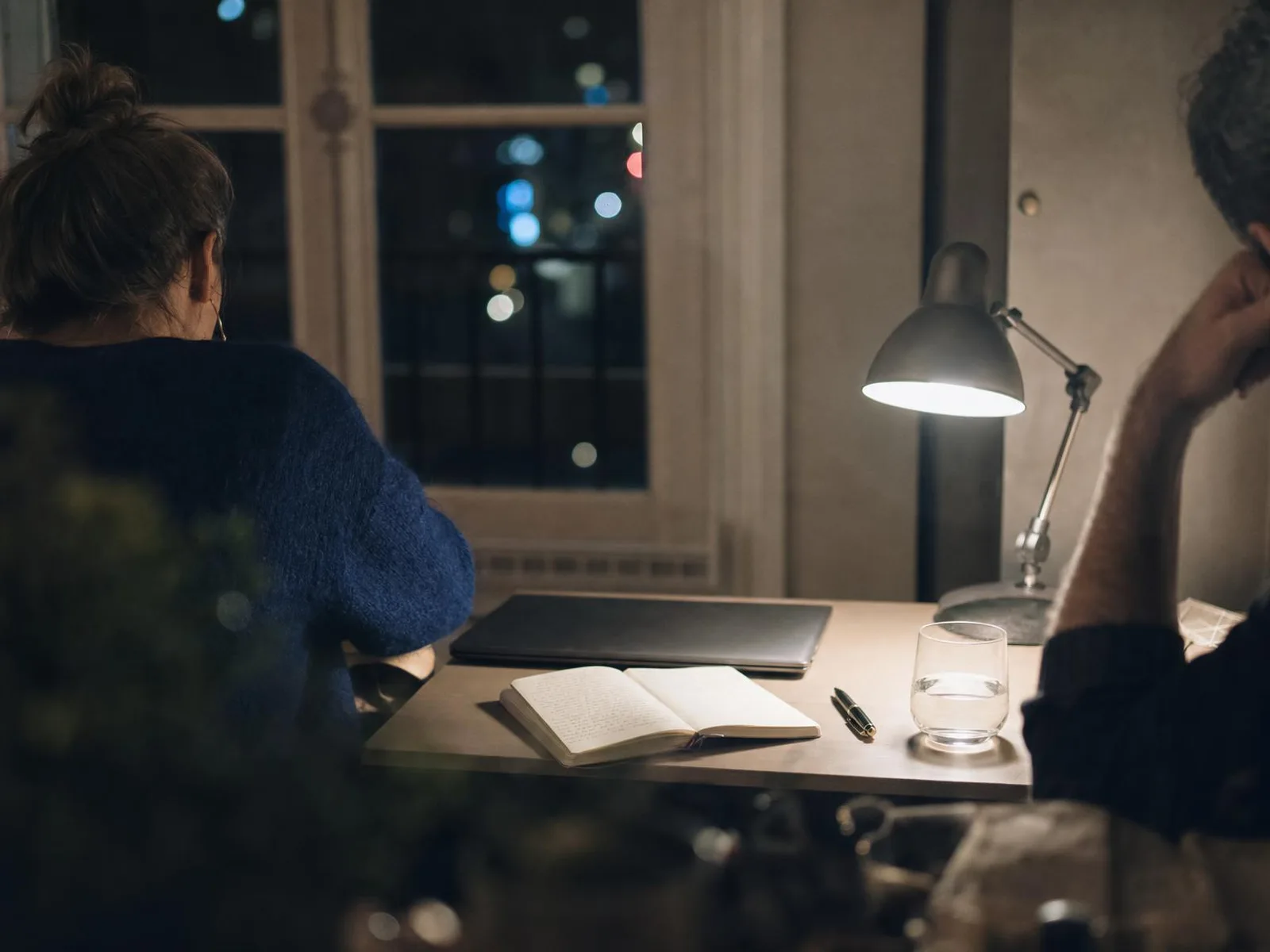 Un bureau d'appartement éclairé par une lampe de bureau, la nuit : un carnet ouvert et couvert d'écriture manuscrite, un stylo posé en travers, un ordinateur fermé et, derrière la fenêtre, des lumières de ville floues.
