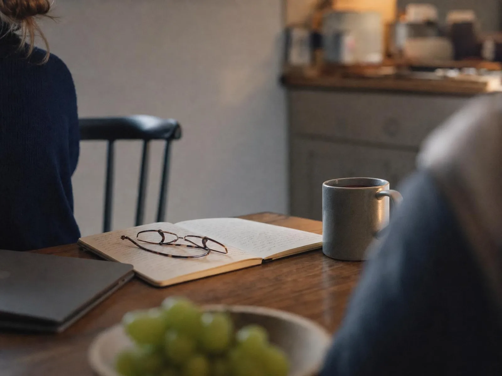 Une table de cuisine le soir, sous une lampe chaude : un carnet ouvert aux pages annotées, une tasse de thé, une paire de lunettes posée sur le papier et un ordinateur fermé sur le côté.