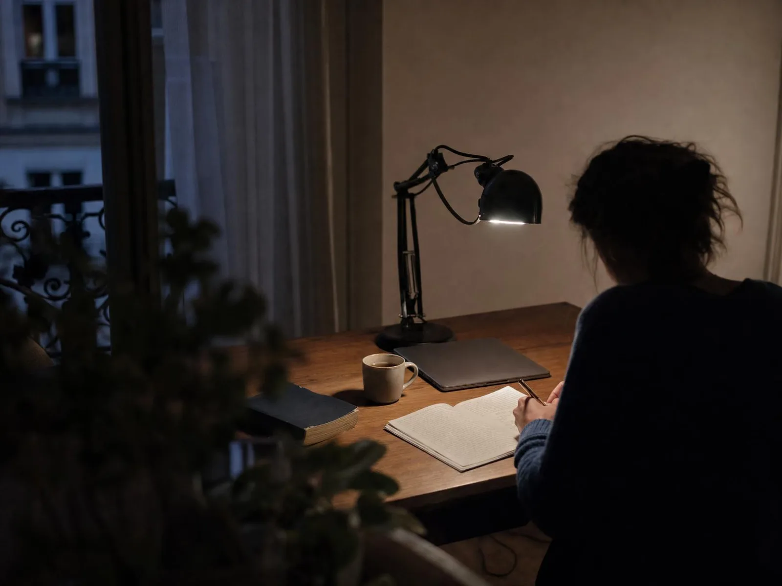 Un bureau éclairé par une lampe seule, la nuit : un cahier ouvert couvert d'écriture, une tasse, un livre de poche posé à l'envers.