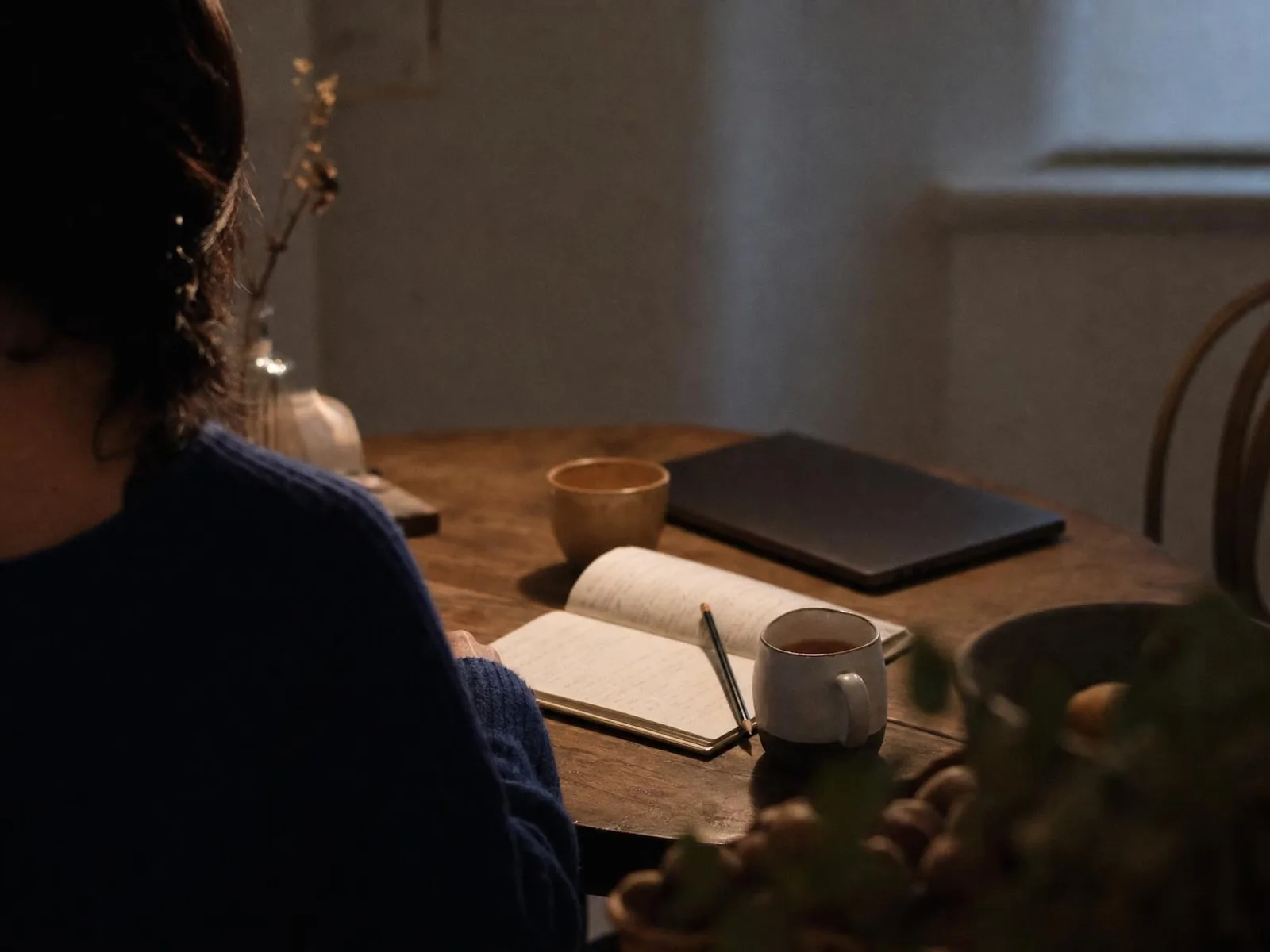 Un cahier ouvert et un crayon posés sur une table de cuisine éclairée par une lampe, une tasse à moitié vide à côté, un ordinateur fermé en arrière-plan.