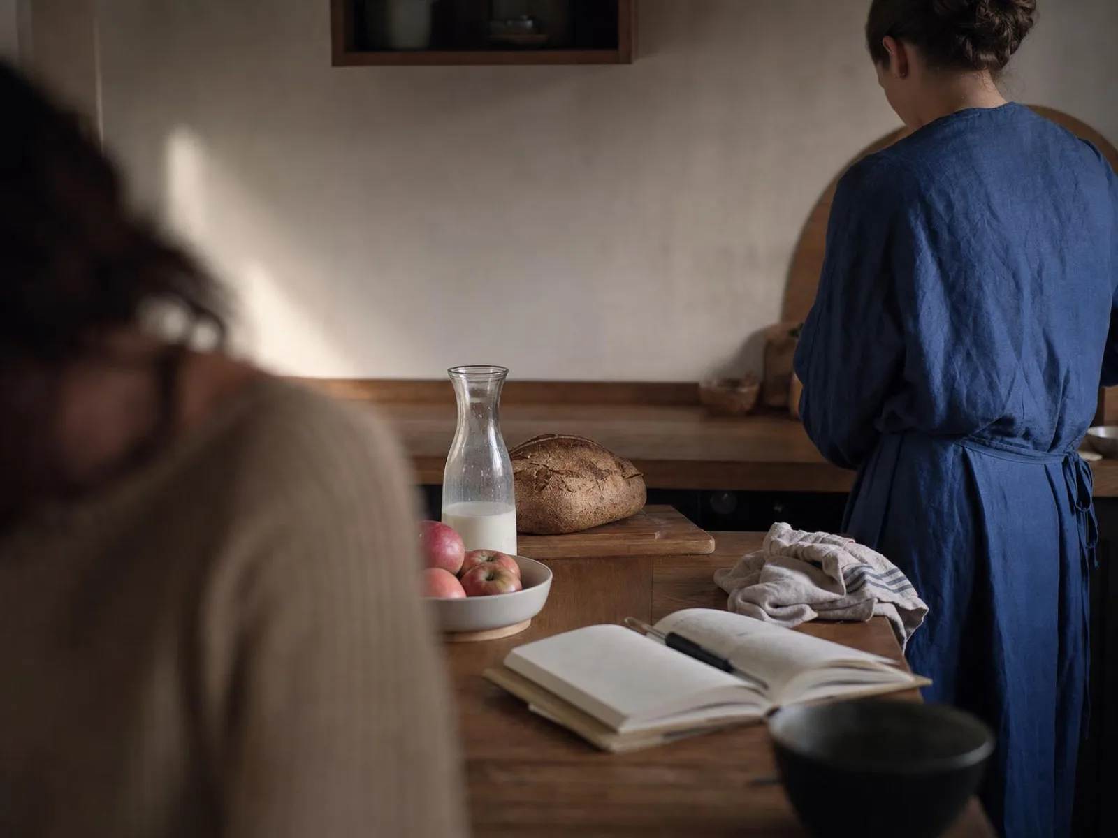 Sur un plan de travail en bois éclairé par une lampe, une carafe de lait, une miche de pain sur une planche, un bol de pommes et un carnet ouvert avec un stylo posé dessus.