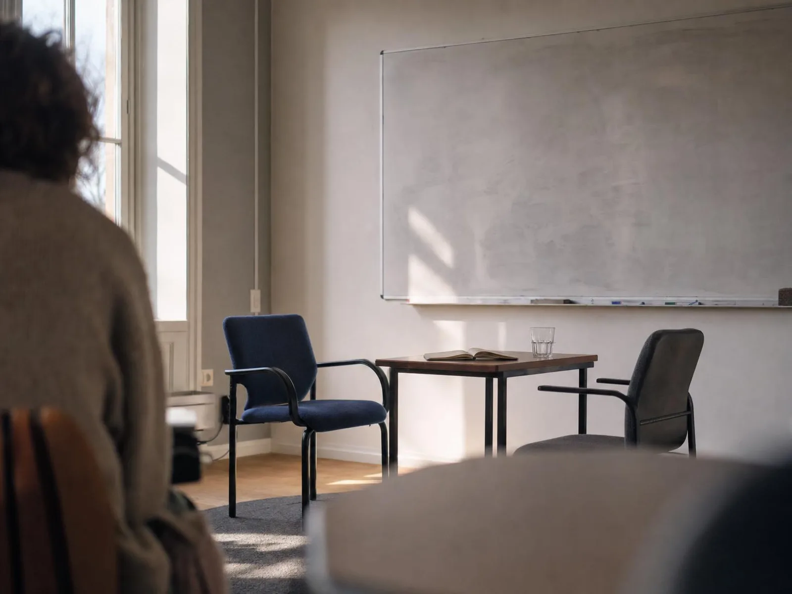 Une salle de cours vide en fin de journée, deux chaises face à face autour d'une petite table, un carnet ouvert et un verre d'eau, la lumière rasante d'une fenêtre haute.