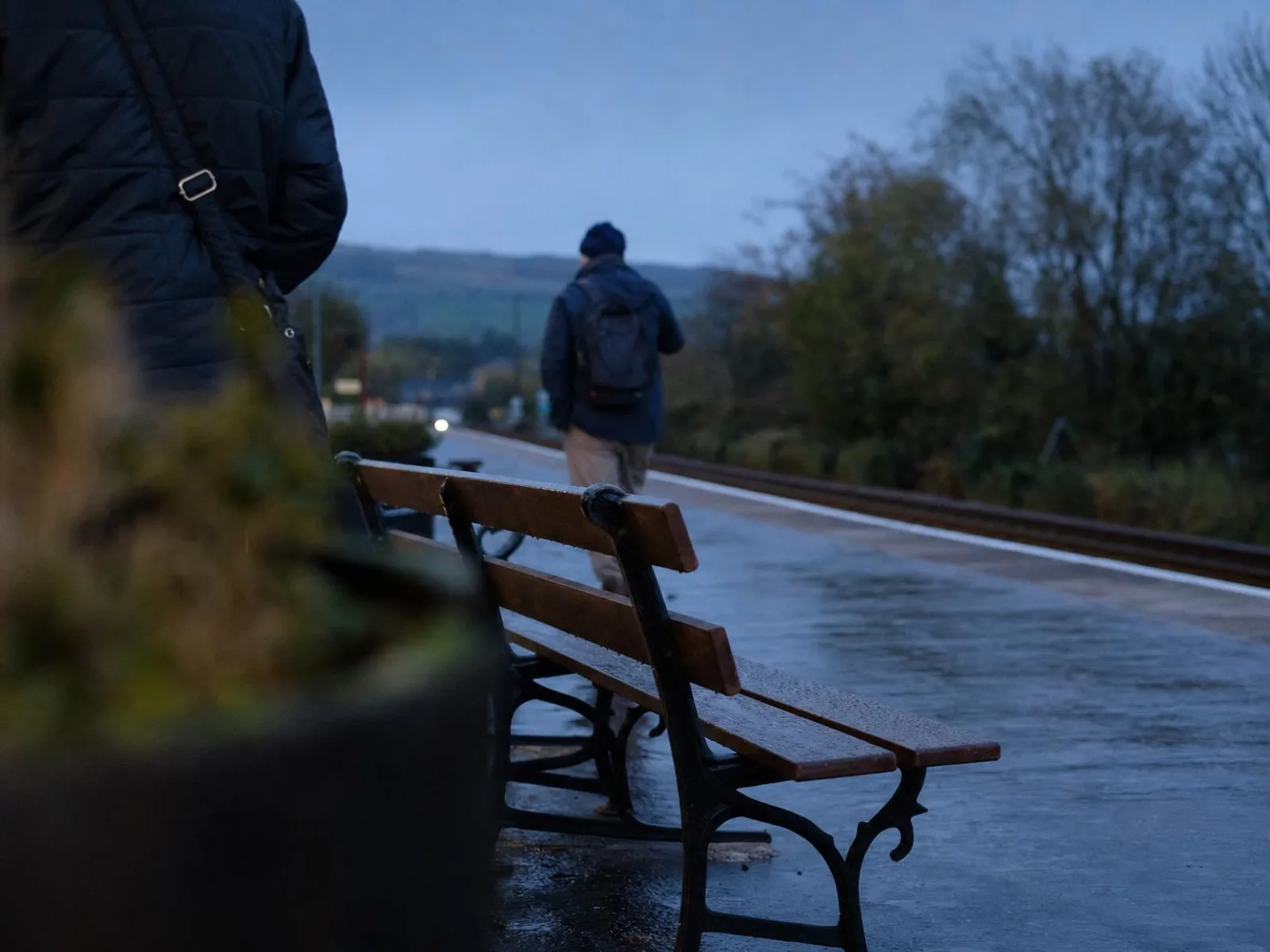 Un quai de gare désert au crépuscule, un banc vide au premier plan et le sol mouillé qui reflète la lumière orangée des lampadaires.
