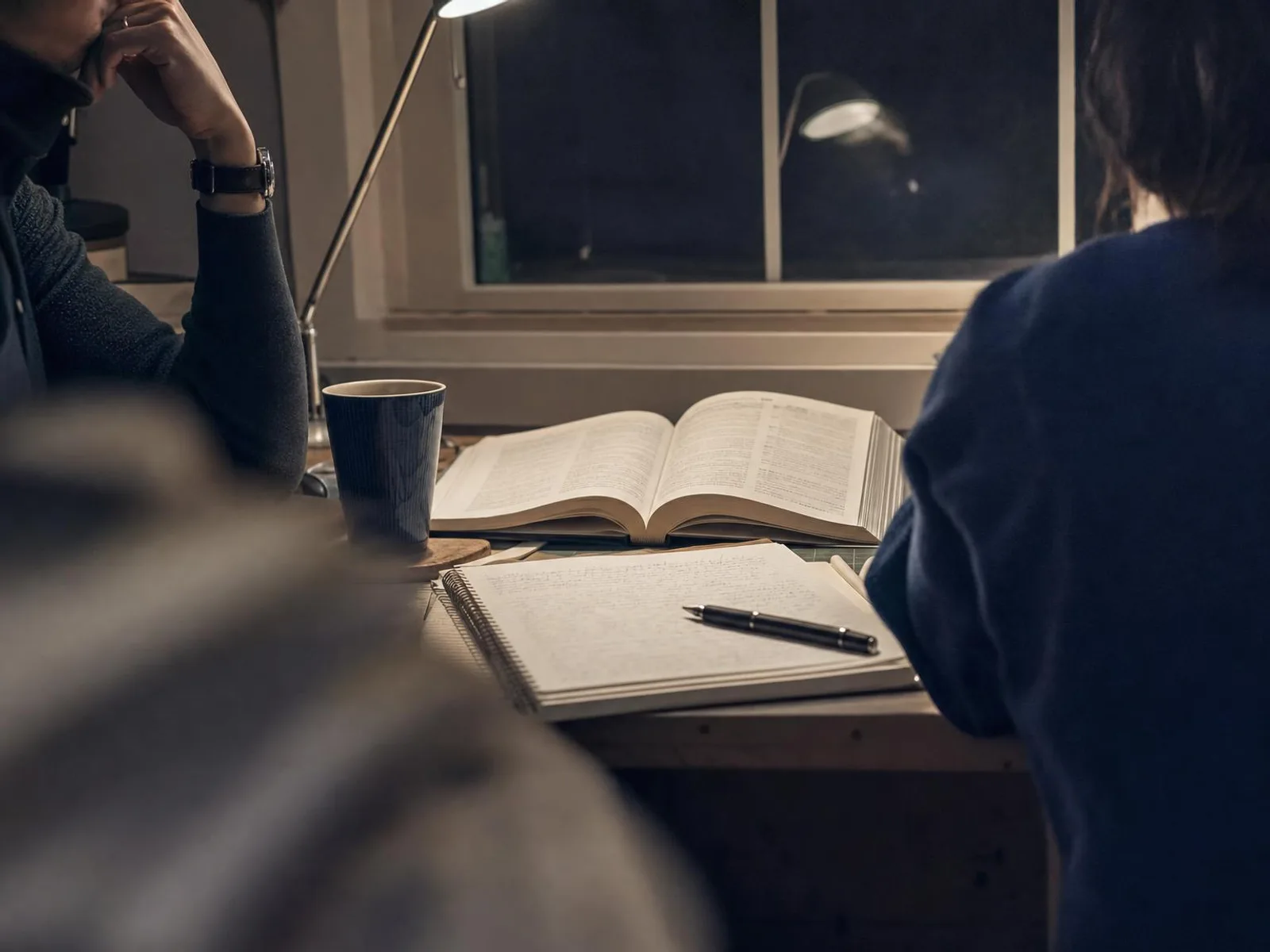 Un bureau de travail éclairé par une lampe la nuit, avec un ouvrage de référence ouvert, un carnet manuscrit, un stylo posé en travers de la page et une tasse de thé.