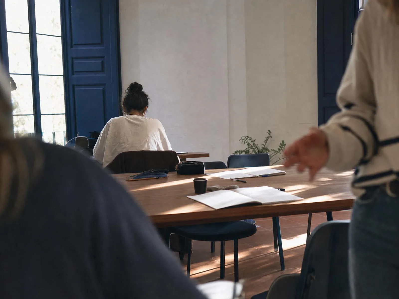 Table de bois près d'une fenêtre à volets, lumière rasante de fin d'après-midi, cahier ouvert aux pages illisibles et tasse de café posée à côté.