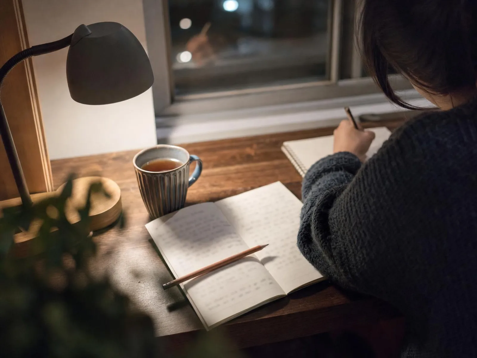 Coin de bureau éclairé par une lampe le soir, avec un carnet ouvert, un crayon posé en travers et une tasse de thé, devant une fenêtre sombre.