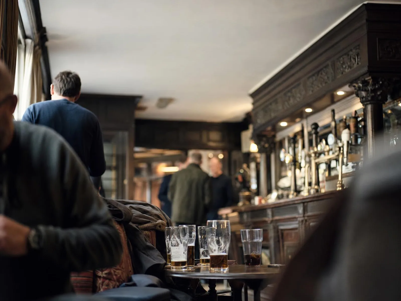 Intérieur d'un pub londonien en début de soirée : boiseries sombres, comptoir en bois et laiton, plusieurs verres posés sur une petite table ronde et des manteaux jetés sur une banquette, silhouettes floues à l'arrière-plan.