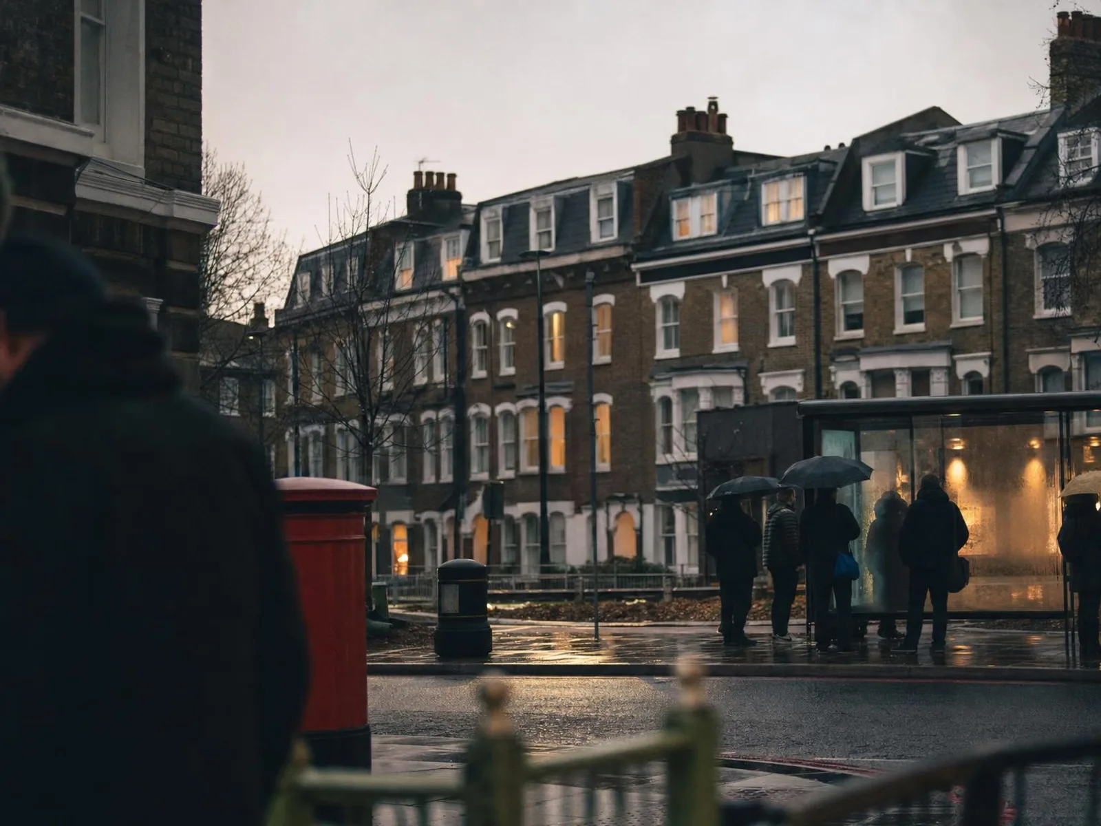 Un coin de rue londonien sous la pluie au crépuscule : boîte aux lettres rouge, trottoir mouillé qui reflète les vitrines, silhouettes floues alignées sous des parapluies à un arrêt de bus, façades de brique en arrière-plan.