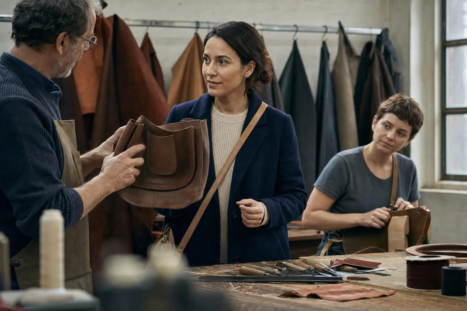 Dans un atelier de maroquinerie, une visiteuse écoute un artisan lui montrer un sac en cours de montage.