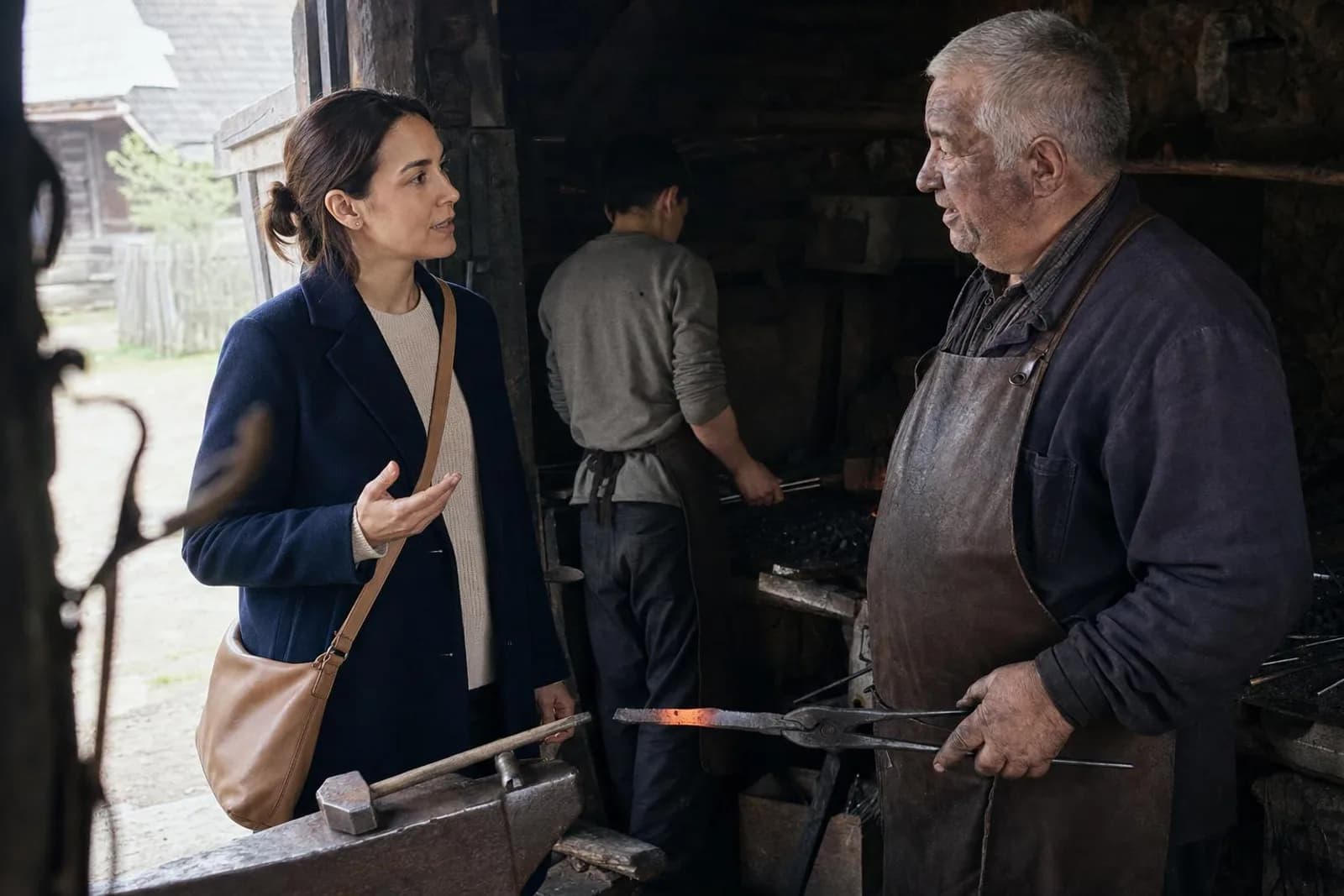 Dans une forge de village, une passante converse avec le forgeron tenant ses tenailles.