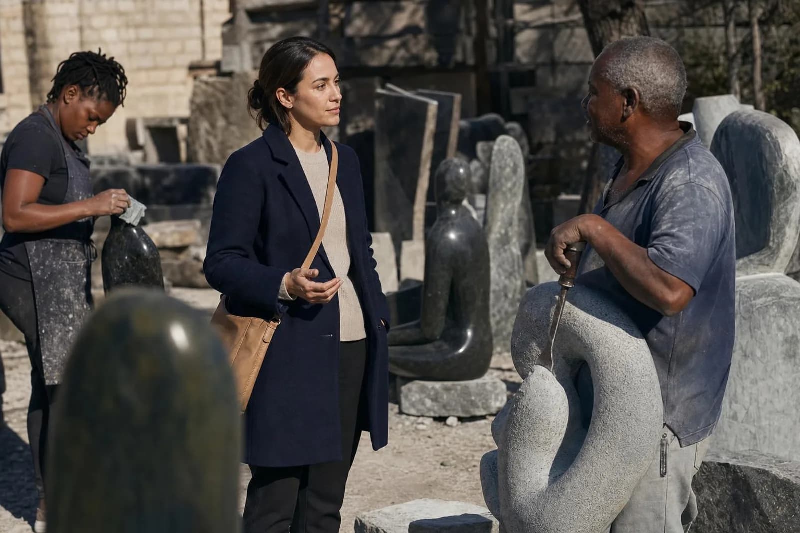 Dans un atelier en plein air, une passante converse avec un sculpteur, ciseau posé contre la pierre.